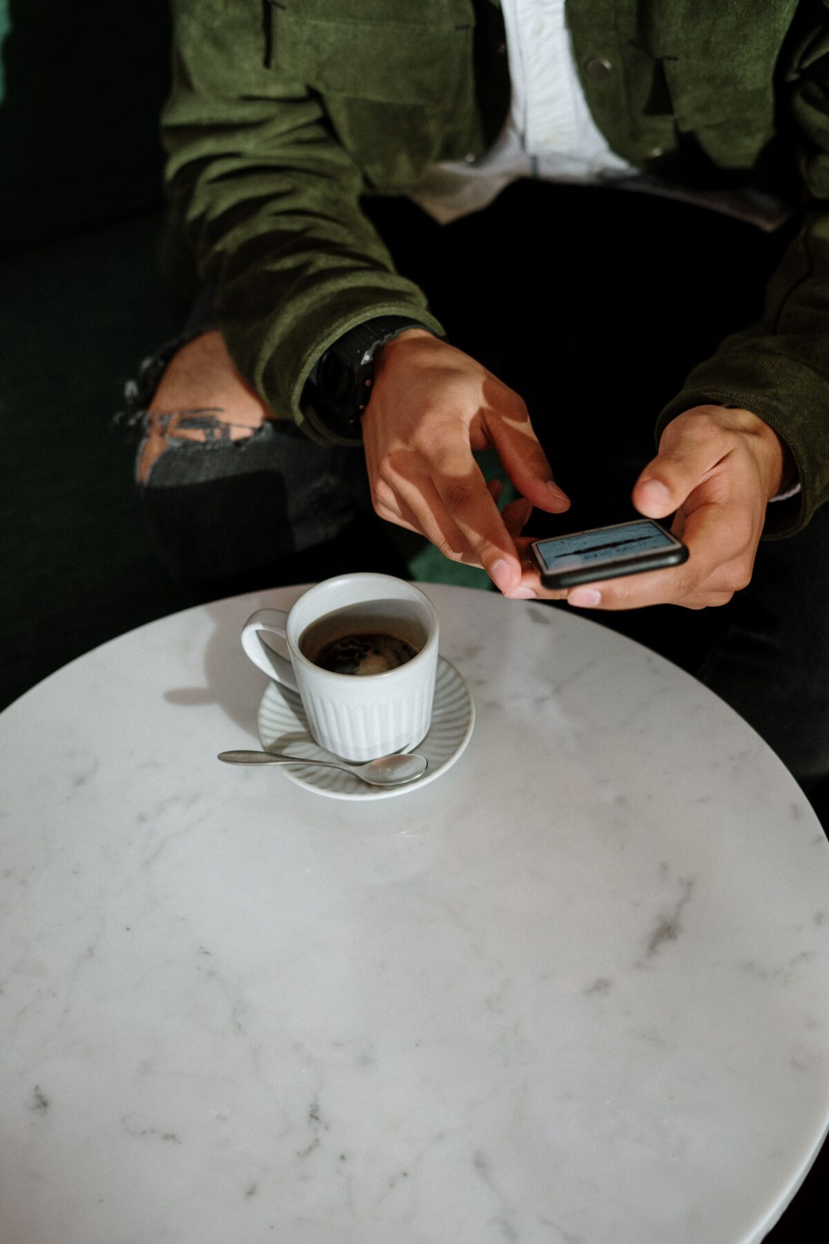 A person wearing a green jacket and ripped jeans is sitting at a white marble table, using a smartphone. There is a cup of coffee with a saucer and spoon on the table. The persons face is not visible.